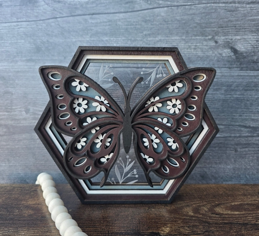 A decorative butterfly-shaped shelf sitter decor piece with layered dark brown wood and white floral inlay, displayed against a gray wood-textured background.