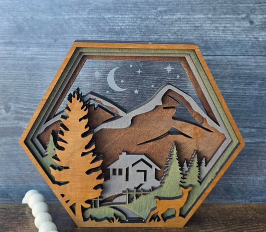 Hexagonal wooden laser-cut wall decor featuring a mountain cabin scene with a crescent moon and stars, displayed on a rustic gray wood background.
