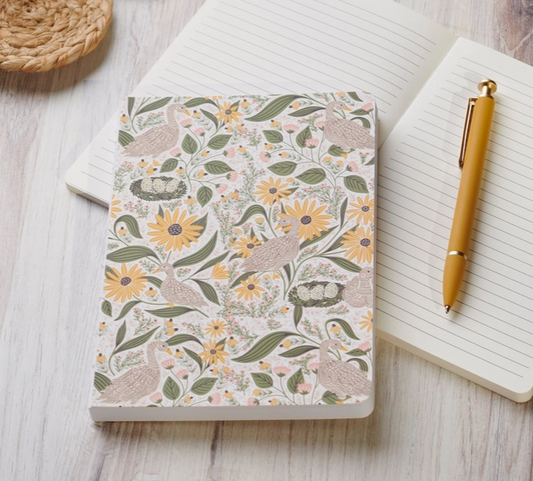 A floral and bird patterned notebook lies on a light wood surface, styled with an open lined notebook and a gold pen.
