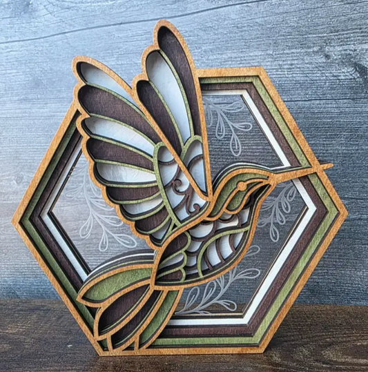 A layered wooden decorative plaque featuring a cutout hummingbird in front of a hexagonal floral frame, photographed on a rustic gray wood surface.
