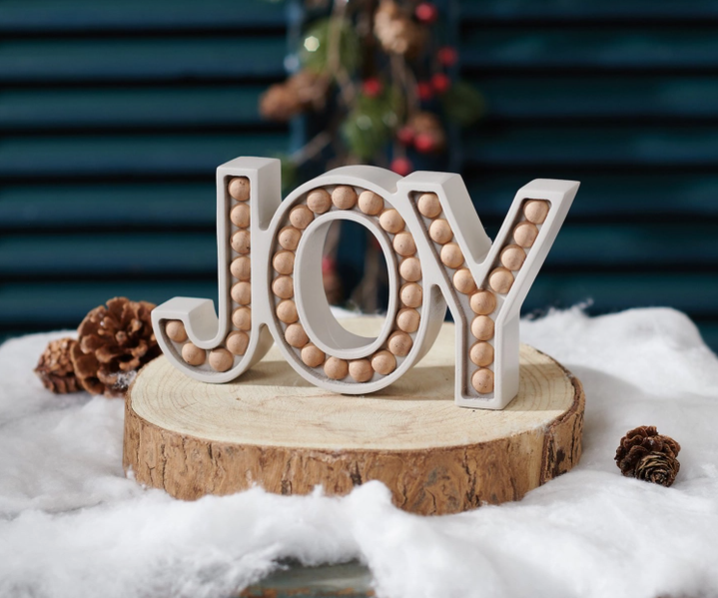 Decorative word 'JOY' made of wooden beads on a wooden base with a Christmas-themed background.