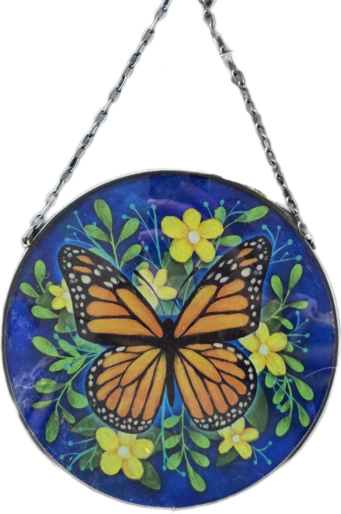 A circular hanging glass art piece featuring a painted monarch butterfly, green leaves, and yellow flowers on a deep blue background, suspended by a black metal chain.