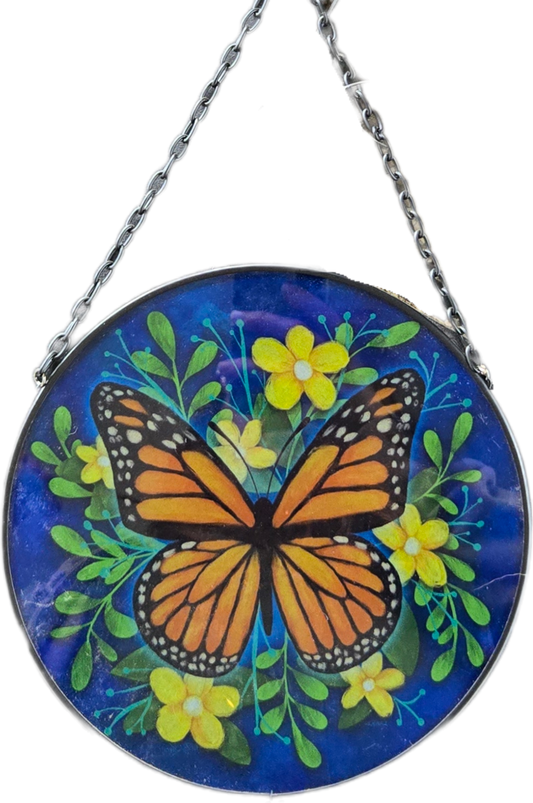 A circular hanging glass art piece featuring a painted monarch butterfly, green leaves, and yellow flowers on a deep blue background, suspended by a black metal chain.