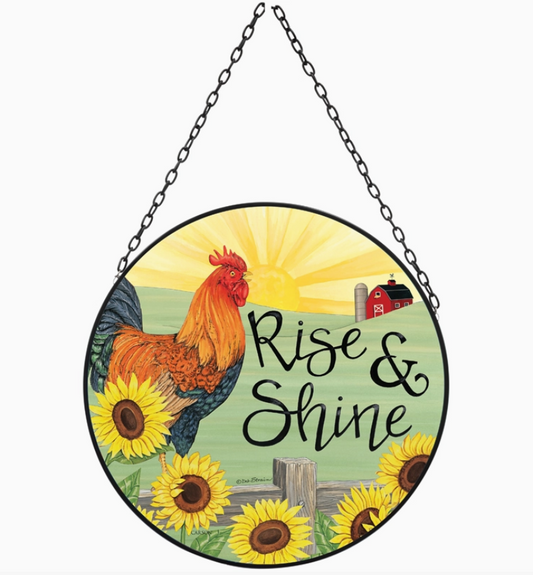 Round hanging sun catcher featuring a colorful rooster, sunflowers, a red barn, and the phrase 'Rise & Shine' in black script, suspended by a black chain against a white background.