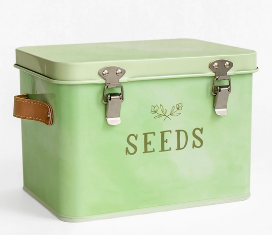 A mint green metal seed storage tin with two silver latch closures, a brown leather handle on the left, and the word 'SEEDS' printed in olive green on the front.