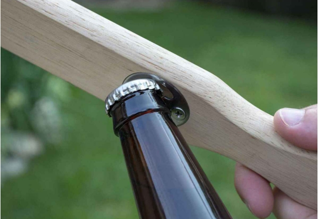 Person holding a brown bottle using a bottle opener on a wooden grill scraper, against a blurred green background